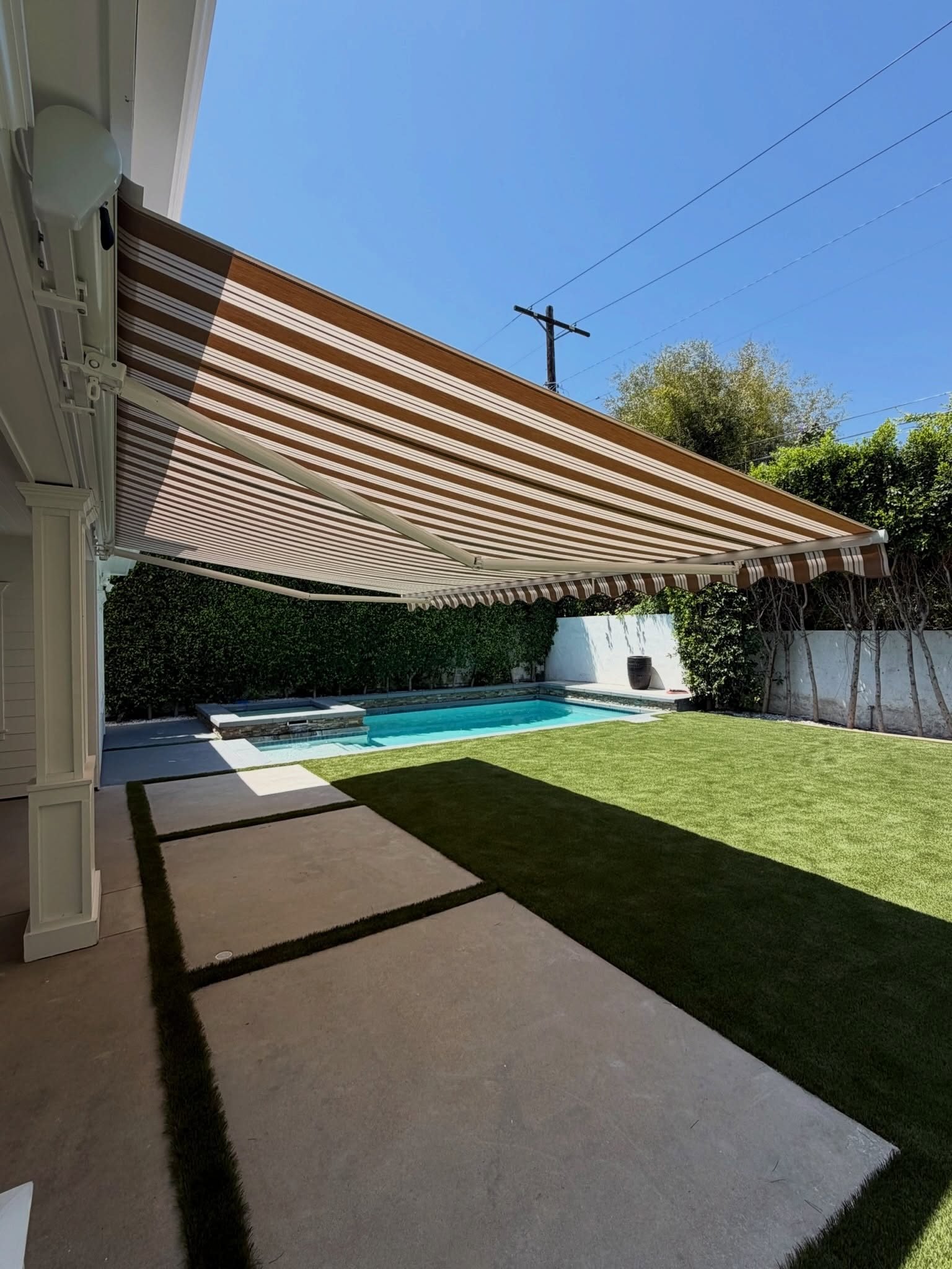 Striped retractable patio awning extending over an outdoor terrace beside a garden and swimming pool